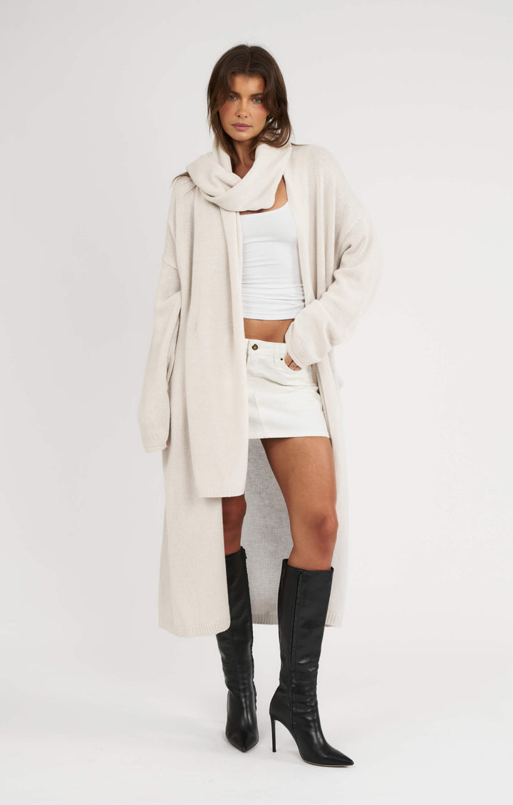 Woman wearing a long beige coat, white top, and black knee-high boots on a white background